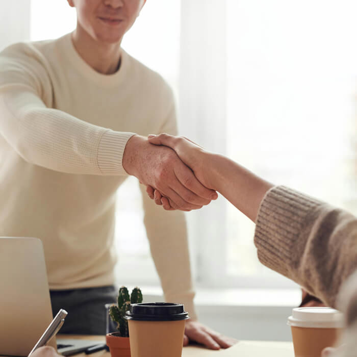A close-up of two individuals shaking hands, one in a light-colored sweater and the other in a darker, textured sweater, set against a softly lit indoor background.