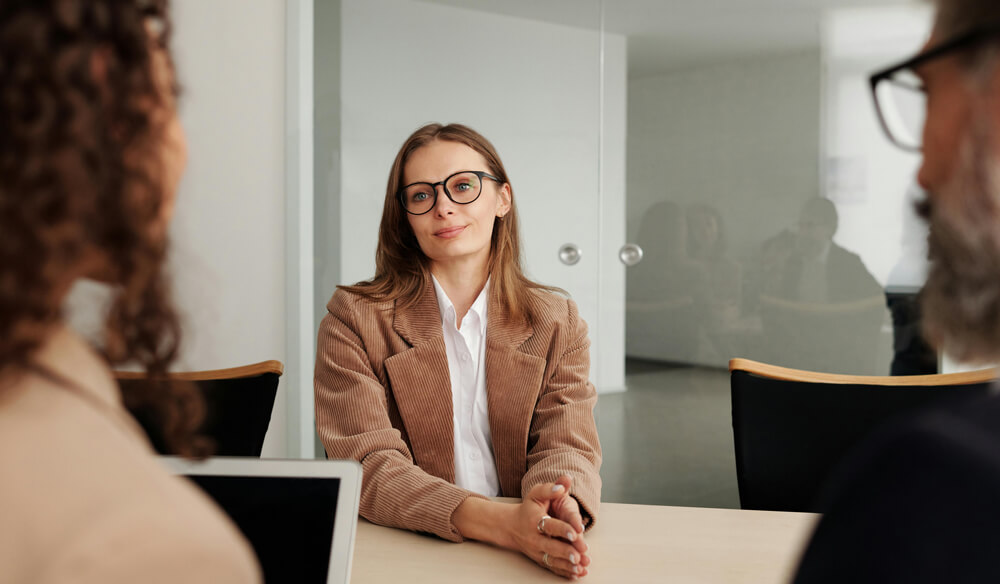 Three individuals engaged in conversation around a table in a modern office setting. The central figure in a brown corduroy blazer sits with hands clasped, while two others—a curly-haired individual and a bearded man with glasses—listen attentively.